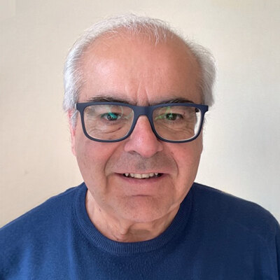 Riccardo, ritratto in primo piano: uomo sorridente con capelli bianchi e occhiali dalla montatura scura, maglia blu, su fondo chiaro